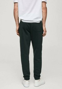 Temno zeleni trenirke iz mehkega materiala, z ravnim krojem in zadnjim žepom, v kombinaciji z belimi supergami.