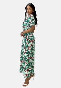 Robe maxi à motif floral avec des nuances de vert et de rouge sur un fond blanc. Dotée de manches courtes bouffantes et d'une taille cintrée.