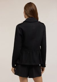 Černý peplum sáčko s límcem, zdobené zlatými knoflíky na ramene a dlouhými rukávy; spárováno s černými šortkami.