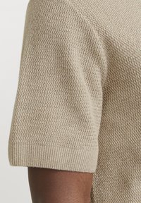 Suéter de punto beige con un patrón texturizado, con mangas cortas y puños acanalados. Tejido suave y ligero visible en primer plano.
