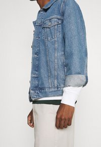 Jeansjacke in Hellblau, ausgestattet mit klassischen Taschen, Knopfschluss und hochgekrempelten Ärmeln über einem in Weiß und Grün geschichteten Hemd.