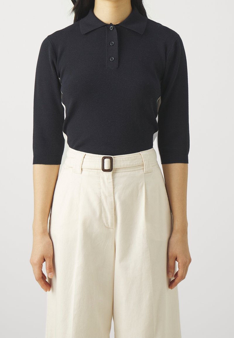 Zwarte poloshirt met lange mouwen en een kraag met een drieknopsinsparing, gedragen met crèmekleurige, hoge taille broeken met een riem.