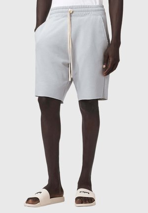 Mężczyzna w jasnoszarych szortach z kieszeniami i białych klapkach z czarnym napisem, stojący na jednolitym tle.