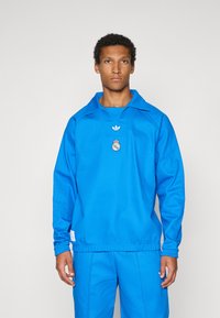 Μπλε αθλητική μπλούζα με μακριά μανίκια και γιακά, με λογότυπο και σήμα στο στήθος. Κατασκευασμένη από λείο ύφασμα με ελαστικά μανίκια και ζώνη στη μέση.