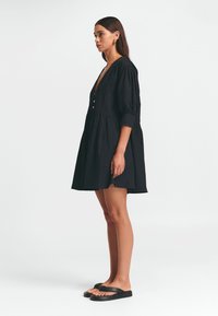 Schwarzes, kurzärmliges Kleid mit tiefem V-Ausschnitt, Knopfdetails und schwingendem Rock. Glatte Stoffqualität, knielang, kombiniert mit schwarzen Sandalen.