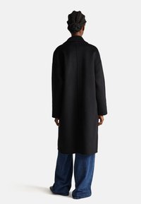 Lungo cappotto nero realizzato in un tessuto liscio, con colletto, spalle cadenti e vestibilità dritta. Indossato sopra pantaloni in denim blu larghi.