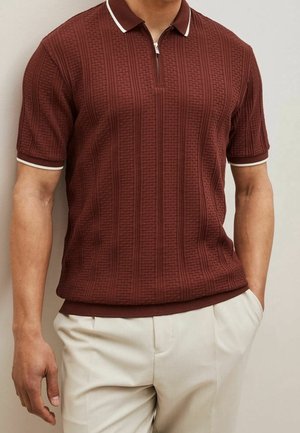 Homme portant un polo à manches courtes texturé bordeaux avec une bordure blanche, associé à un pantalon beige clair, debout devant un fond uni.