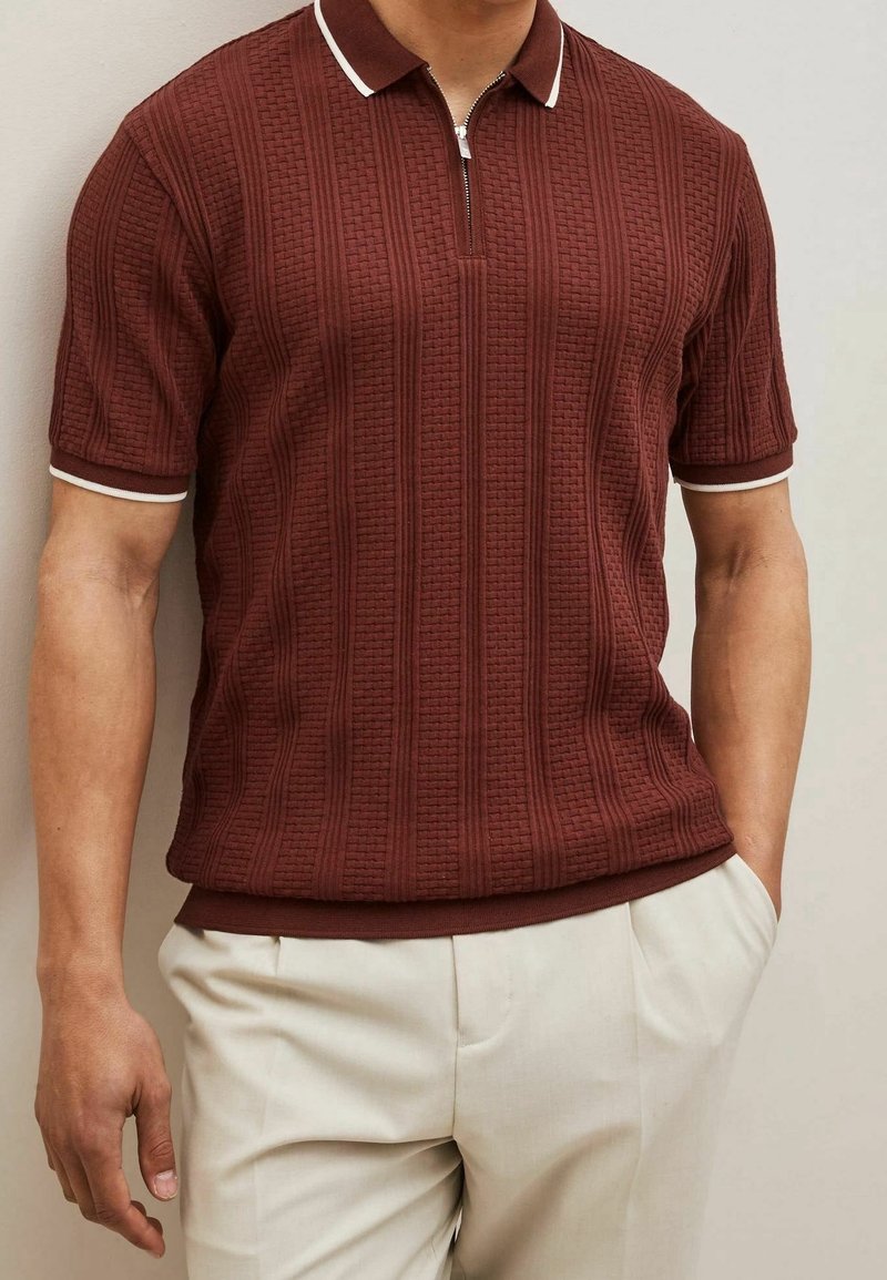 Homme portant un polo à manches courtes texturé bordeaux avec une bordure blanche, associé à un pantalon beige clair, debout devant un fond uni.