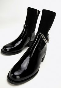 Czarne lakierowane botki do kostki z czarną tkaniną na cholewce, zapięcie na zamek i srebrny łańcuszek na kostce. Niski, masywny obcas.