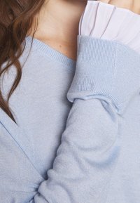 Ljusblå stickad tröja med ribbad halsringning och muddar. Har en lager av vit, plisserad tygdekoration vid handleden. Mjukt material.