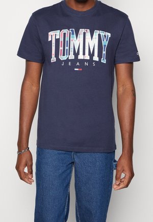 Mies, joka pitää yllään laivastonsinisen Tommy Jeansin t-paidan, jossa on iso monivärinen "TOMMY" teksti, sekä farkut, seisoo rentoine käsivarsineen.
