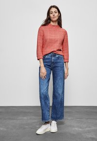 Maglione a maglia rosso con texture, collo alto, maniche a tre quarti; jeans a gamba larga blu; sneakers bianche; sfondo grigio uniforme.