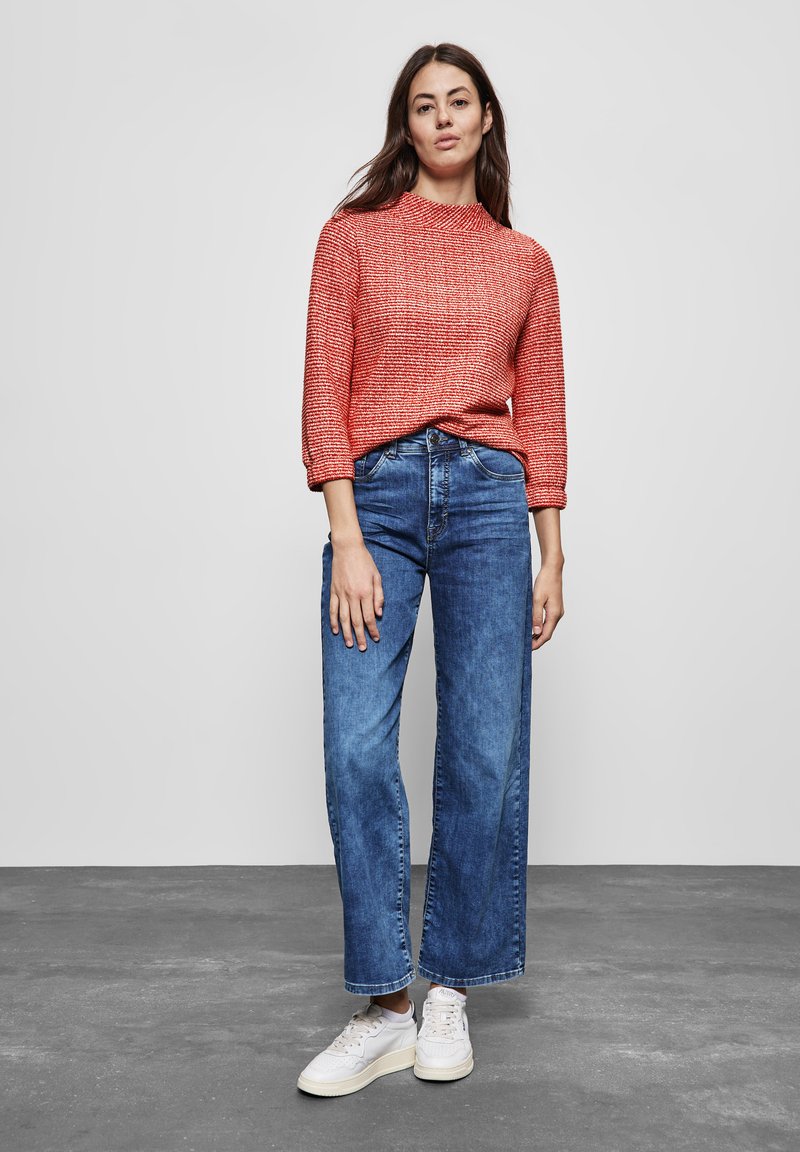 Maglione a maglia rosso con texture, collo alto, maniche a tre quarti; jeans a gamba larga blu; sneakers bianche; sfondo grigio uniforme.