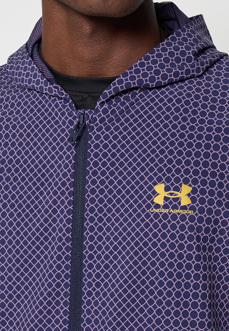 Giacca a vento navy con cappuccio, caratterizzata da un motivo geometrico a cerchi rosa. Presenta una zip frontale e un logo Under Armour giallo sul petto.