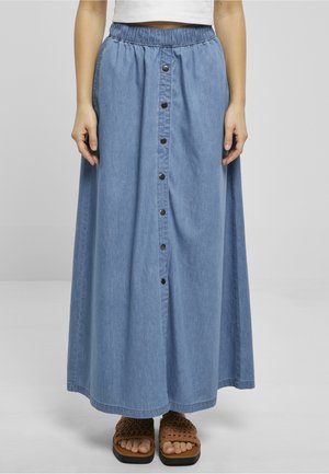 Mujer que lleva una falda vaquera larga azul con botones delanteros y cintura elástica, combinada con sandalias marrones tejidas y un top corto blanco.