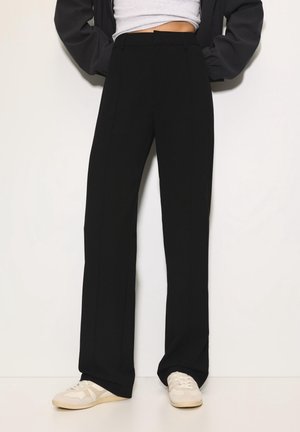 Person trägt schwarze, hoch taillierte, weit geschnittene Hose, weiße Turnschuhe, weißes, kurzes Oberteil und schwarze Jacke mit Händen in den Taschen, steht vor einer weißen Wand.