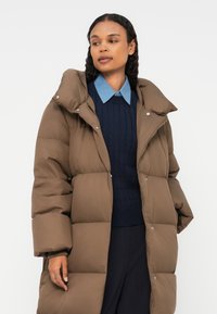 Hnědý zateplený kabát s vysokým límcem, velkými nadýchanými částmi a zapínáním na patentky. Nese se přes tmavě modrý svetr s světle modrou košilí.