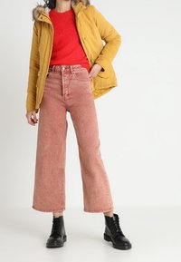 Żółta kurtka puchowa z futrzanym kapturem, czerwona teksturowana sweter, oraz różowe, szerokie jeansy z wysokim stanem. Czarne botki na sznurówki.