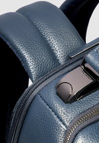 Mochila de cuero azul con un acabado texturizado, correas acolchadas, una cremallera suave y un detalle de herrajes metálicos. El diseño presenta formas estructuradas.