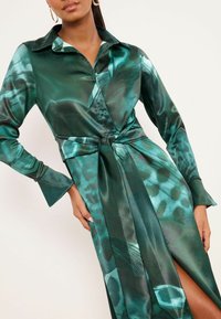 Rochie verde din satin, cu nasturi în față, mâneci lungi și talie cu curea, având un model abstract bogat și o crăpătură înaltă până la coapsă.