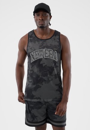 Man draagt een zwart camouflagemesh hemd met de tekst "NEW ERA" en bijpassende shorts, zwarte pet en zilveren kettingen, staand tegen een grijze achtergrond.