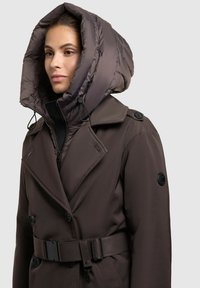 Donna che indossa un cappotto marrone scuro con cintura e un ampio cappuccio imbottito, in piedi contro uno sfondo grigio chiaro uniforme.