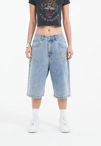 Shorts de mezclilla de color azul claro con un corte relajado, hasta la rodilla, que presentan un detalle de logo bordado en el muslo izquierdo. Se llevan calcetines blancos y zapatillas.