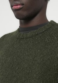Pull vert en tricot avec une finition texturée, un col rond, et une chemise noire superposée en dessous. Vue rapprochée du col et du tissu.