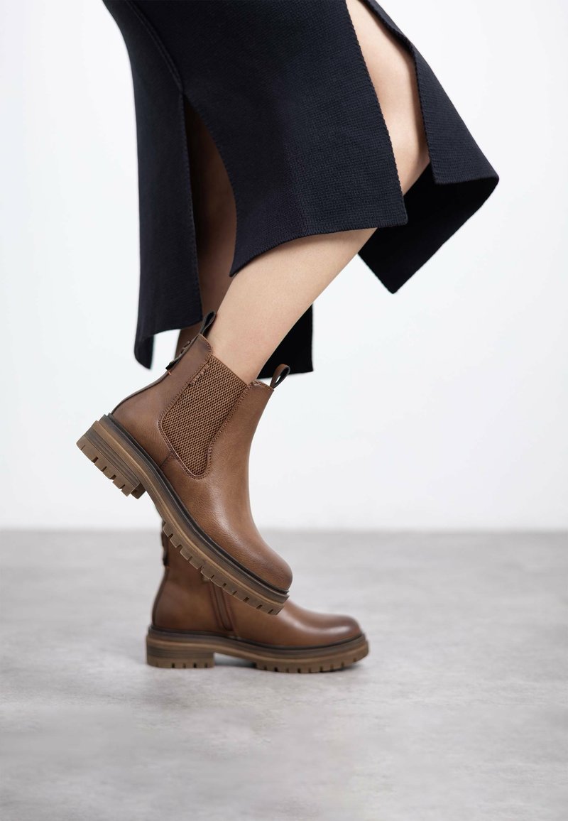 Botas de tobillo de cuero marrón con paneles laterales elásticos, superficie texturizada, suelas resistentes y con tacos, y tiradores. Llevadas con una falda de punto negra.