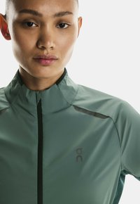 Jacheta sport ușoară verde, cu guler înalt, fermoar frontal și accente negre. Prezintă un logo pe pieptul stâng. Textură netedă.