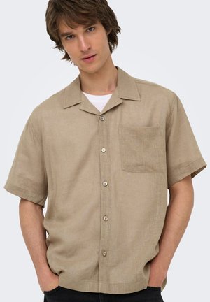 Jeune homme portant une chemise beige à manches courtes avec poche poitrine et un sous-t-shirt blanc, mains dans les poches, fond gris clair neutre.