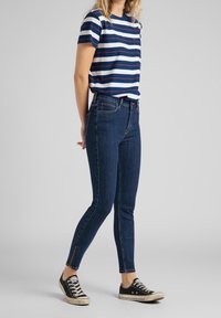 Camiseta de rayas azul y blanca de mangas cortas, combinada con jeans ajustados azul oscuro con cremalleras en los tobillos y zapatillas negras.