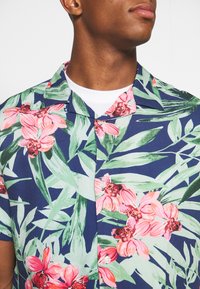 Kurzärmeliges Button-up-Shirt mit navy-blauem Hintergrund und großen pinken Blumen- sowie grünen Blattmustern. Aus leichtem Stoff gefertigt.