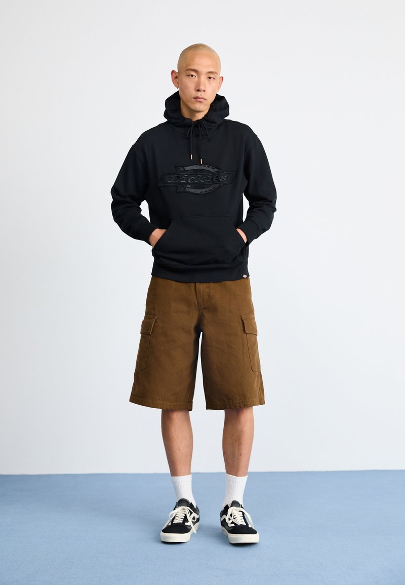 Felpa nera con un logo testurizzato, abbinata a pantaloni cargo marroni. Calzini bianchi alla caviglia e sneakers nere completano l'outfit. Sfondo neutro.