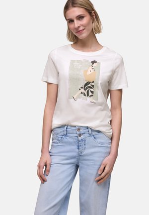 Frau trägt weißes T-Shirt mit Grafik einer gehenden Frau, die eine beige Tragetasche trägt, kombiniert mit hellblauen Jeans.