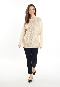 Beige pulóver elől flitterekkel, hosszú ujjakkal és kerek nyakkal, fekete, slim-fit nadrággal és fekete, hegyes orrú cipővel párosítva.