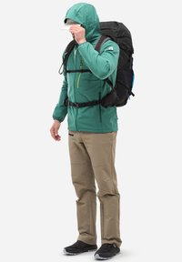 Grüne Kapuzenjacke mit einem schwarzen Rucksack, beigen Hosen und schwarzen Schuhen. Die Jacke verfügt über eine Reißverschlusstasche und verstellbare Ärmelbündchen.