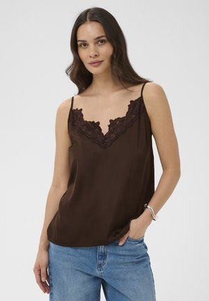 Femeie purtând un maiou maro închis cu dantelă și blugi albastru deschis, pozând cu o mână în buzunar pe un fundal simplu.