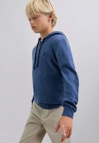 Sudadera azul con capucha hecha de tela suave con dobladillo y puños acanalados. Presenta un pequeño logo de calavera en el pecho. Se lleva con pantalones caqui.