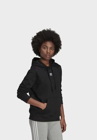 Felpa nera con cappuccio regolabile e tasca a marsupio, con il logo Adidas bianco sul petto. Abbinata a pantaloni della tuta grigi.