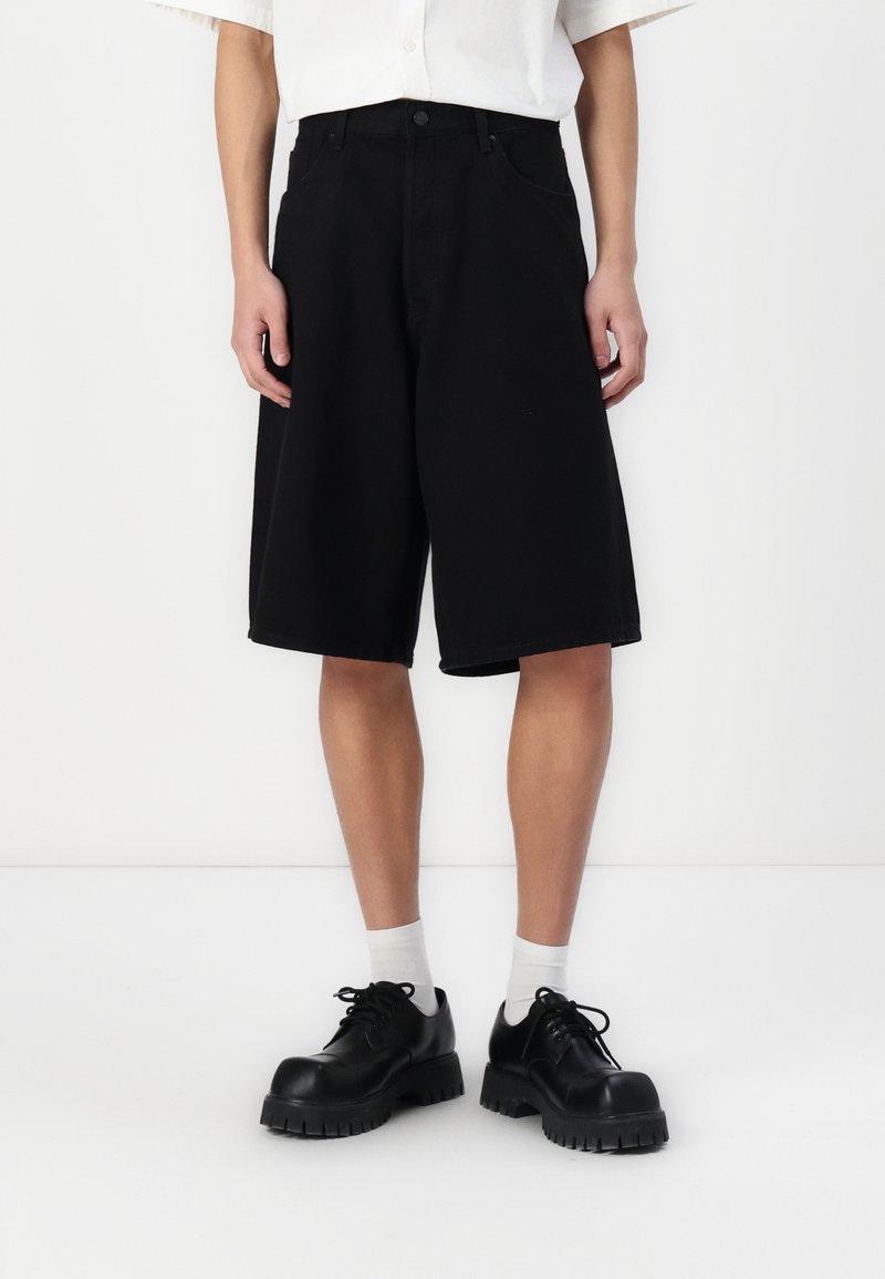 Shorts de mezclilla negra con un corte holgado, que cuentan con bolsillos delanteros y cremallera, combinados con zapatos negros de cordones y calcetines blancos.