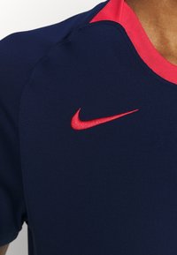 Marinblå idrottströja med en rosa krage, som pryds av en röd Nike-logotyp på bröstet. Materialet verkar slätt och elastiskt.