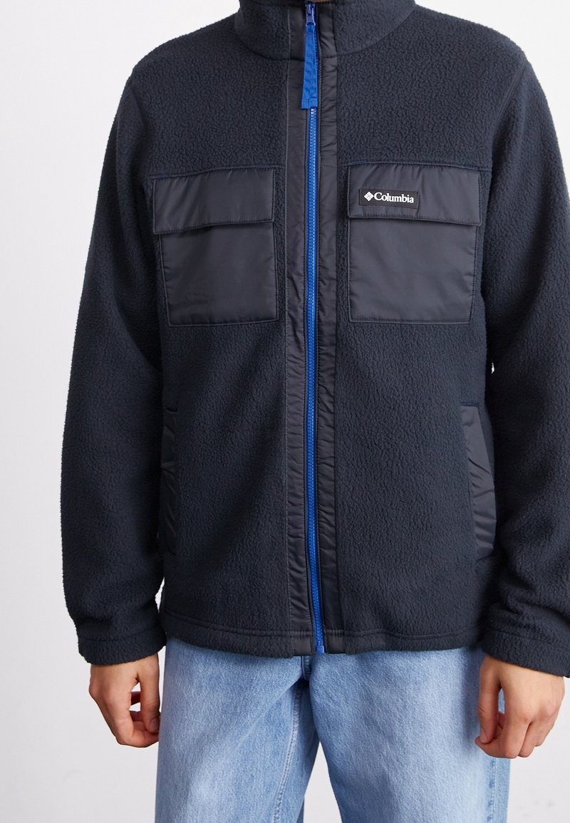 Czarna bluza polarowa z zamkiem błyskawicznym z przodu, niebieskim suwakiem i dwoma przednimi kieszeniami z patkami; wyróżnia się teksturowaną tkaniną i miękkim kołnierzem.