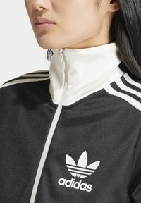 Černá atletická bunda na zip s bílým límcem a třemi bílými pruhy na ramenou. Na hrudi je vyražené logo adidas.