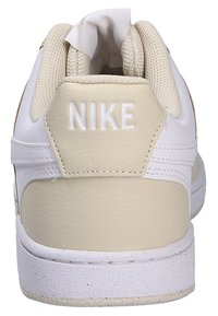 Zapatilla blanca con acentos beige, de cuero texturizado y malla, con un logotipo "NIKE" en relieve en el talón y una suela de goma.