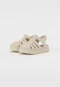 Beige rubber sandals with a closed toe, striped cutouts, and back strap. Soft texture with a lightweight, chunky sole design.