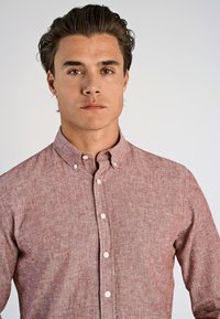 Ung man med mörkt hår som bär en knappad, långärmad, ljusröd linne skjorta, som står mot en enkel, ljus bakgrund.