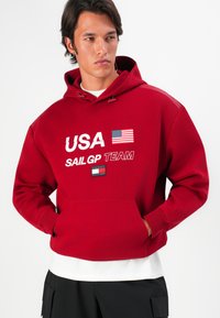 Rød hoodie med teksten "USA" og "SAIL GP TEAM", grafik af det amerikanske flag, kængurulomme og snoretræk i hætten med ribbede manchetter.