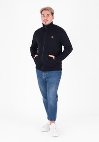 Maglione zip-up nero con colletto alto, polsini a coste e due tasche frontali, abbinato a jeans blue e sneakers bianche.