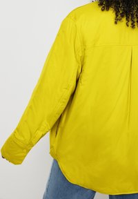 Žlutá vyp paddingová bunda s volným střihem, má zádový sklady, boční kapsy a manžety na knoflíky. Hladká textura a neformální design.